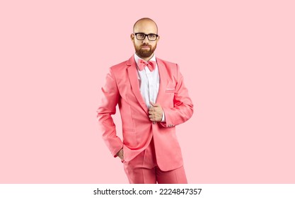 Studio shot of handsome bald young man in pink suit. Confident guy with ginger beard in fashionable suit, bowtie and trendy glasses standing with hand in pocket isolated on solid pink color background