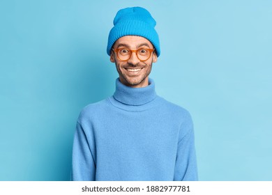 Studio shot of handsome cheerful guy has positive face expression looks through optical glasses wears hat and turtleneck isolated over blue background. Male teenager in stylish clothes poses indoor - Shutterstock ID 1882977781