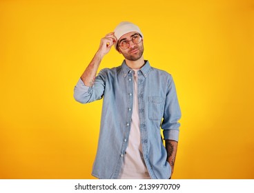 Studio Shot Of  Handsome Man In Jeans , Glasses And White Hat On Yellow Background. Thinking Concept. Tatoo And Beard. 