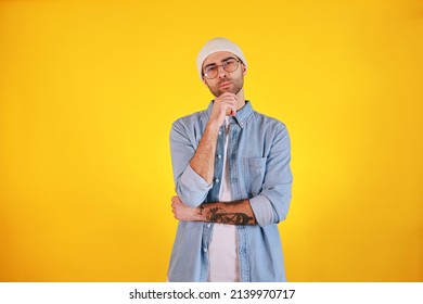 Studio Shot Of  Handsome Man In Jeans , Glasses And White Hat On Yellow Background. Thinking Concept. Tatoo And Beard. 