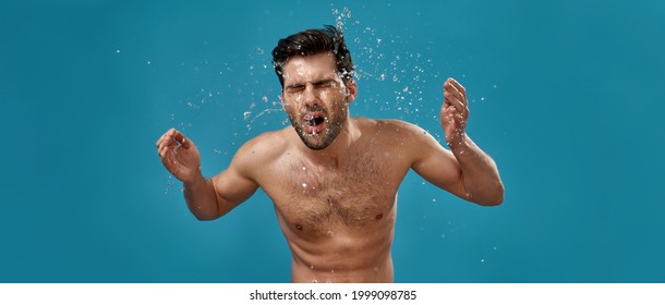 Studio shot of handsome naked man washing his face with many water splashes around isolated over blue background. Skin care and cleaning concept. Web Banner