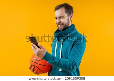 Studio shot of handsome smiling bearded young man holding basketball ball in hand, watching play live broadcast online and making bets at favourite team using bookmaker's mobile application.