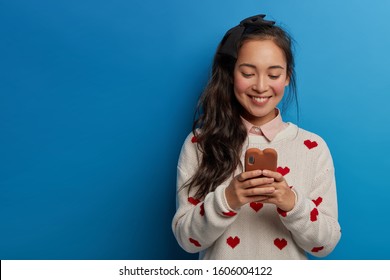 Studio Shot Of Happy Asian Woman With Pony Tail Holds Smartphone, Dressed In Casual Clothes, Types Messages, Uses Interesting App, Smiles Happily, Isolated Over Blue Wall. People, Technology Concept