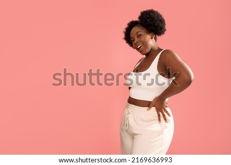 Studio shot of happy dark skinned, young woman with afro hairstyle, smiling and posing over pink pastel studio background. Real people emotions concept. Body positive. Copy space. 
