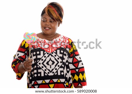 Studio shot of happy fat black African woman smiling while holding and looking at heart shaped lollipop