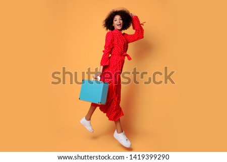 Studio shot of happy playful black woman  fooling around and jumping on orange background. Holding blue   suitcase. Red trendy dress. Tourism and holiday concept