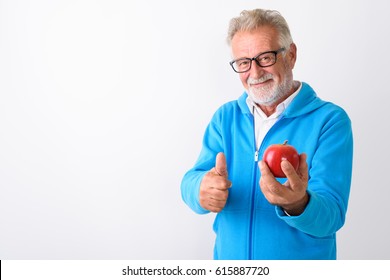 Studio Shot Of Happy Senior Bearded Man Smiling While Holding Red Apple And Giving Thumb Up Ready For Gym Against White Background