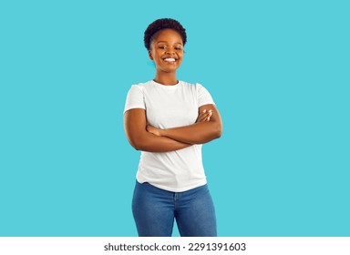 Studio shot of a happy, smiling woman in casual clothes. Portrait of a beautiful young African American girl in a white T shirt and jeans standing with her arms crossed isolated on a blue background