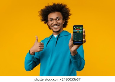 Studio shot of happy smiling young man showing thumbs up and reaching smartphone with bookmaker's application interface on its screen. Sport bet, money win concept. Focus is in phone.