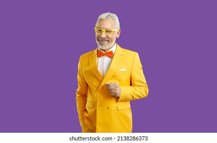 Studio shot of happy white haired bearded senior man wearing bright yellow suit, white shirt, orange bow tie and trendy glasses standing with his hand in pocket isolated on solid purple background - Powered by Shutterstock