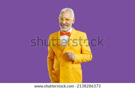 Studio shot of happy white haired bearded senior man wearing bright yellow suit, white shirt, orange bow tie and trendy glasses standing with his hand in pocket isolated on solid purple background