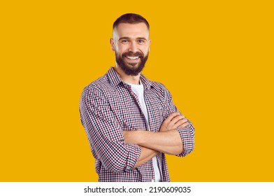 Studio shot of a happy young guy. Cheerful handsome bearded young man wearing a casual shirt standing with his arms folded isolated on a yellow color background, looking at the camera and smiling