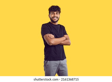 Studio Shot Of Happy Young Man In Casual Wear. Handsome Indian Guy With Modern Haircut In Black Tshirt Standing With His Arms Folded Isolated On Yellow Background, Looking At Camera And Smiling