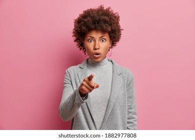 Studio shot of impressed curly haired woman has eyes full of disbelief points index finger at camera notices shocking thing dressed grey formal clothes isolated over pink background. Omg concept
