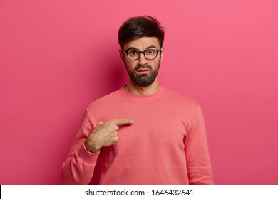 Studio shot of indignant man points at himself, feels embarrassed and displeased, points at himself, asks whether he is guilty, wears casual clothes, poses against rosy studio wall. Who, me?