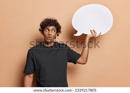 Studio shot of isolated on beige background young handsome hindu guy with black curly short hair wearing dark blue tshirt holding white big speechbubble at his raised right hand and looking at it