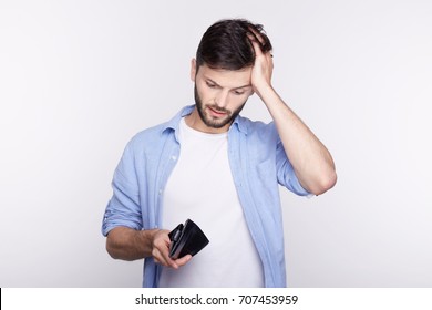 Studio shot isolated on white wall. Young bearded man dressed casual  found his  stylish black leather wallet almost empty. The crisis hits financial well-being of handsome Caucasian office manager.


