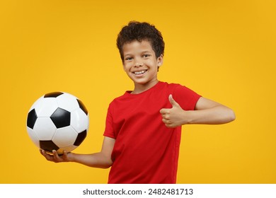 Studio shot of joyful African American schoolboy wearing red t-shirt, posing over yellow background holding the ball in hand and showing thumbs up gesture.                                - Powered by Shutterstock