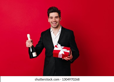 Studio Shot Of Man With Champagne And Gift Isolated On Red. Smiling Guy In Suit Holding Bottle Of Wine