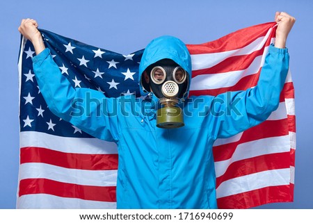Studio shot of man ecologist holding USA flag behind him, wearing uniform and gas mask, posing isolated over blue background, scientist tries to prevent exhausting toxic gases into air. Ecology.