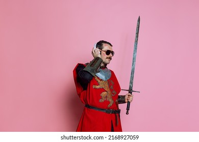 Studio Shot Of Man, Medieval Warrior Or Knight Listening To Music In Headphones With Delightful Expression Isolated Over Pink Background. Comparison Of Eras, History, Funny Meme Emotions