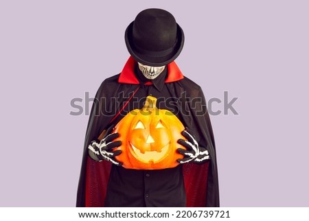 Studio shot of man in skeleton costume holding jack-o-lantern. Man wearing black hat, black and red cloak and gloves standing isolated on light purple background and holding orange Halloween pumpkin