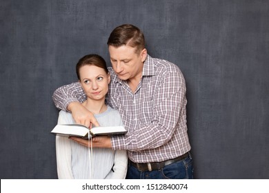 Studio Shot Of Man Trying To Pay Attention Of Displeased Woman To Text In Book While She Is Bored And Tired Of Reading And Tutoring, Being Not Interested In Tedious Studying, Over Gray Background