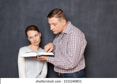 Studio Shot Of Man Trying To Pay Attention Of Displeased Woman To Text In Book While She Is Bored And Tired Of Reading And Studying, Being Not Interested In Tedious Activity, Over Gray Background