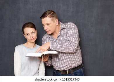 Studio Shot Of Man Trying To Pay Attention Of Annoyed Woman To Interesting Info In Book While She Is Bored And Tired Of Reading, Being Not Interested In Tedious Learning, Over Gray Background