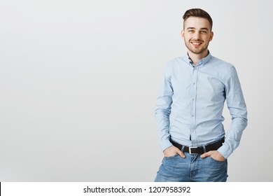 Studio shot of neat stylish handsome young male entrepreneur in blue collar shirt and jeans holding hands in pockets and smiling with optimistic friendly smile preparing for audition or interview