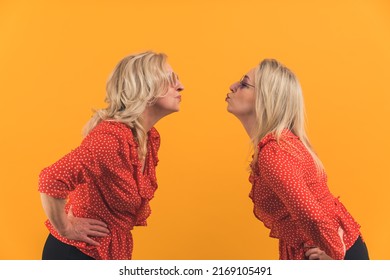 Studio Shot On Yellow Background Of Two Cute Millenial Blonde Women Standing Apart And Giving Kisses To Each Other From Distance. High Quality Photo