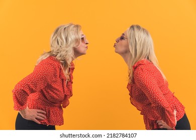 Studio Shot On Yellow Background Of Two Cute Millenial Blonde Women Standing Apart And Giving Kisses To Each Other From Distance. High Quality Photo