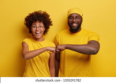 Studio Shot Of Overjoyed Satisfied Woman And Man With Dark Skin, Give Fist Bump, Agree To Work Together, Have Success In Project Work, Wear Yellow Attire, Pose In Studio. Cooperation Concept