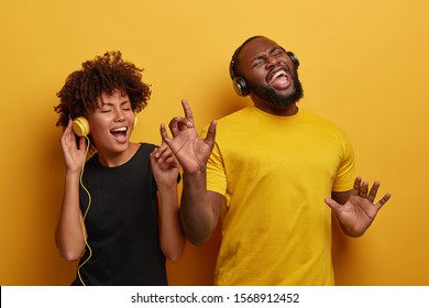 Studio shot of overjoyed teenage couple dance with raised hands, enjoy cool music in stereo headphones, listen pleasant popular track, move actively against yellow background, feel happy and great