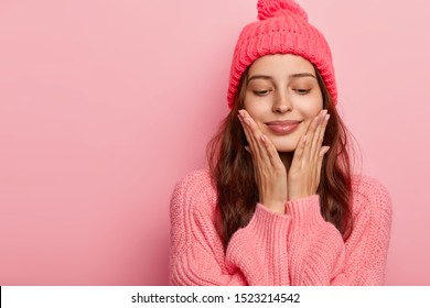 Studio Shot Of Pleasant Looking Female Model Touches Chin With Both Palms, Has Eyes Closed, Enjoys Delicate Well Cared Skin, Has Dark Hair, Dressed In Winter Outfit, Isolated On Pink Wall, Blank Space