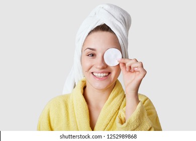 Studio shot of pleasant looking young smiling European woman covers eye with cotton disk, has white towel on head, takes care of skin, models against white studio wall. Beauty procedure at home