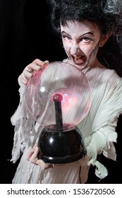 Studio Shot Portrait Of Young Girl In Costume Dressed As A Halloween, Cosplay Of Scary Bride Of Frankenstein Posing With Glass Lightning Ball