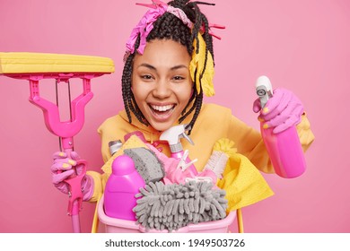 Studio Shot Of Positive Dark Skinned Woman Cleans Apartment Smiles Gladfully Holds Mop And Cleaning Detergent Satisfied With Results Isolated Over Pink Background. Housework And Chores Concept