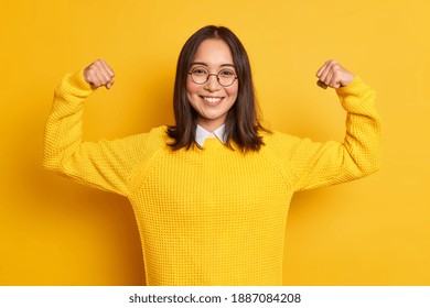 Studio shot of positive young Asian woman raises arms shows muscles pretends to be very strong and powerful smiles gently wears casual sweater isolated over yellow background. Look at my biceps