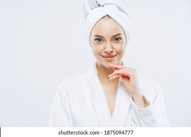 Studio shot of pretty beauty woman has washed hair, wears wrapped towel on head, has manicure, cute natural face, touches chin gently, looks with tender smile, dressed in bath robe, poses indoor - Powered by Shutterstock