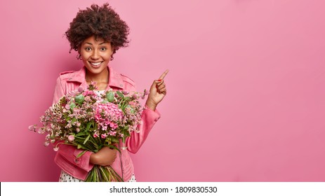 Studio shot of pretty dark skinned woman with bunch of beautiful flowers, shows direction, points on copy space against pink background, celebrates birthday, has festive mood. Bouquet, blossom