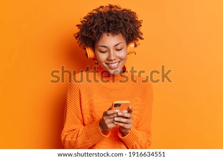 Studio shot of pretty dark skinned woman chooses song from playlist uses smartphone and wireless headphones dressed in long sleeved jumper isolated over orange background. Technology lifestyle