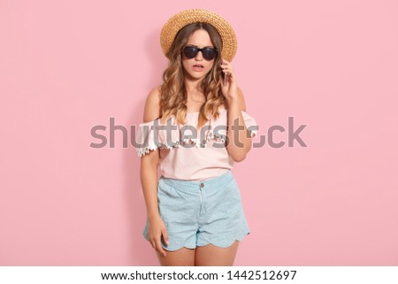 Studio shot of pretty woman wearing blouse, short, straw hat and sunglasses, having conversation via smart phone, model posing isolated over pink studio background. People and technology concept.