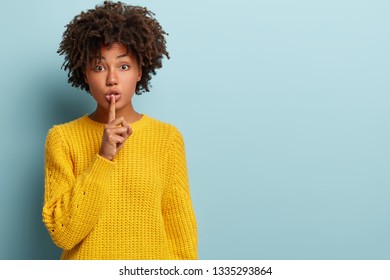 Studio shot of quiet dark skinned lady keeps fore finger over lips, makes silence gesture, gossips with friend, wears yellow sweater, says hush, isolated over blue studio wall with blank space - Shutterstock ID 1335293864