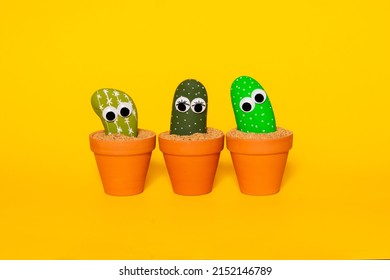 Studio shot of rocks painted to look like cactus and given plastic googly eyes to animate them arranged in a small group in terra cotta pots sitting on sand on a seamless white background. - Shutterstock ID 2152146789