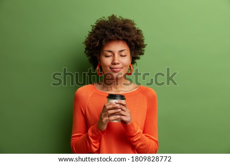 Studio shot of satisfied dark skinned girl with Afro hair holds paper cup of hot coffee, closes eyes, wears orange jumper, green background. Beautiful young woman enjoys aromatic cappuccino.