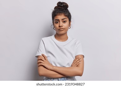Studio shot of serious Indian woman with dark hair combed in bun keeps arms folded waits for explanations listens attentively information dressed in basic t shirt isolated over white background.
