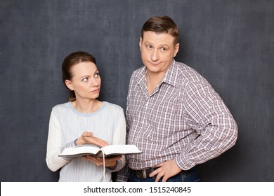 Studio Shot Of Serious Woman Trying To Pay Attention Of Displeased Man To Important Info In Book While He Is Bored And Tired Of Reading, Being Not Interested In Tedious Activity, Over Gray Background