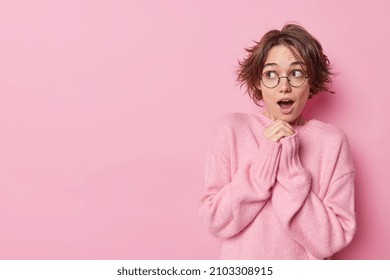 Studio Shot Of Shocked Young Woman Looks Away With Widely Opened Mouth Keeps Hands Together Feels Astonished Concentrated Away Wears Cashmere Jumper Isolated Over Pink Background Copy Space.