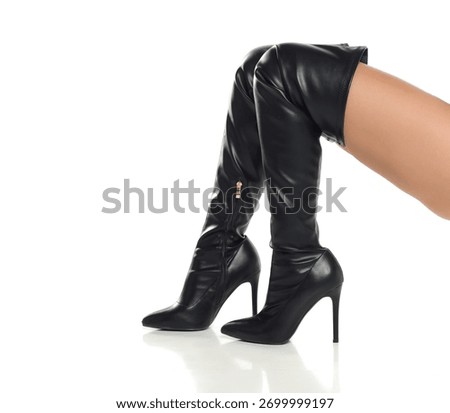 Studio shot, side angle view of a woman’s legs in black high boots and a black bodysuit kneeling on a white background